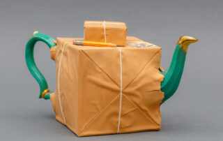 A ceramic teapot that appears as if the green and gold object is wrapped and sticking out of brown paper and rope. A smaller ceramic package and pencil sit atop the main portion.