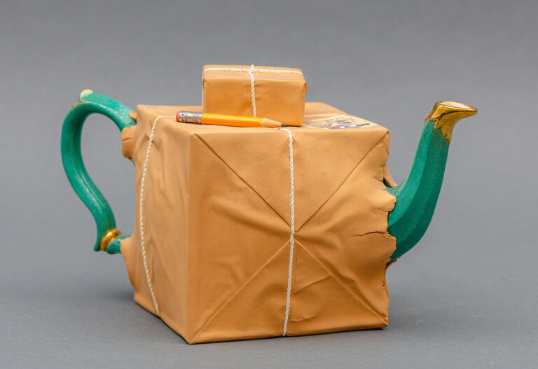 A ceramic teapot that appears as if the green and gold object is wrapped and sticking out of brown paper and rope. A smaller ceramic package and pencil sit atop the main portion.