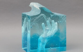 A dense blue glass sculpture. The exterior is squared except for the top, which is cut to resemble a wave. An open-palm hand can be seen in the interior as it reaches towards the top of the piece.