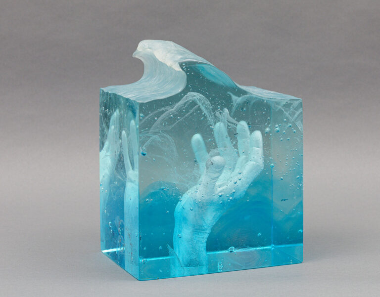 A dense blue glass sculpture. The exterior is squared except for the top, which is cut to resemble a wave. An open-palm hand can be seen in the interior as it reaches towards the top of the piece.