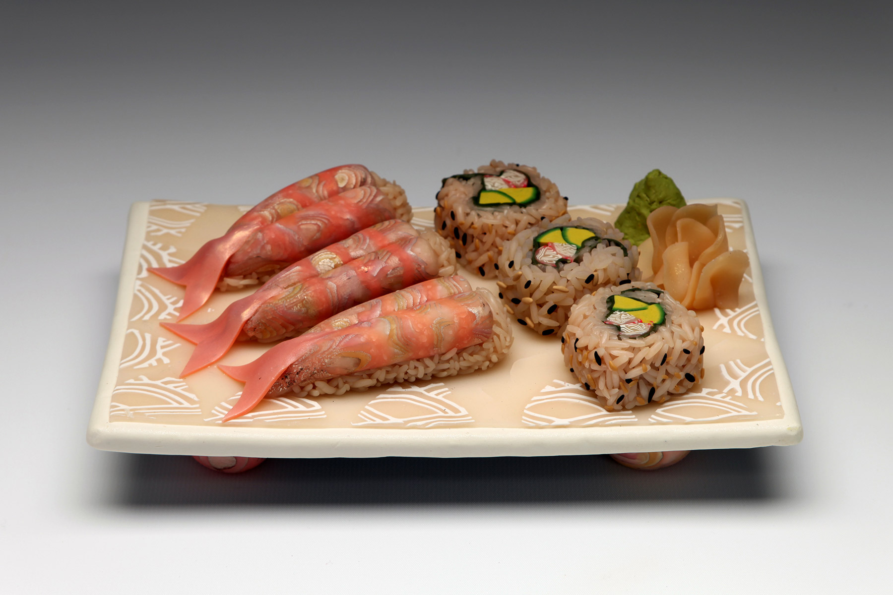 Polymer clay formed into the shape of a light tan/pink decorated platter. Atop the platter are three pieces of faux sashimi on the left with brown rice and pink fish; three pieces of faux sushi in the center with brown rice and green and pink filling; and faux ginger on the right with faux wasabi behind it.