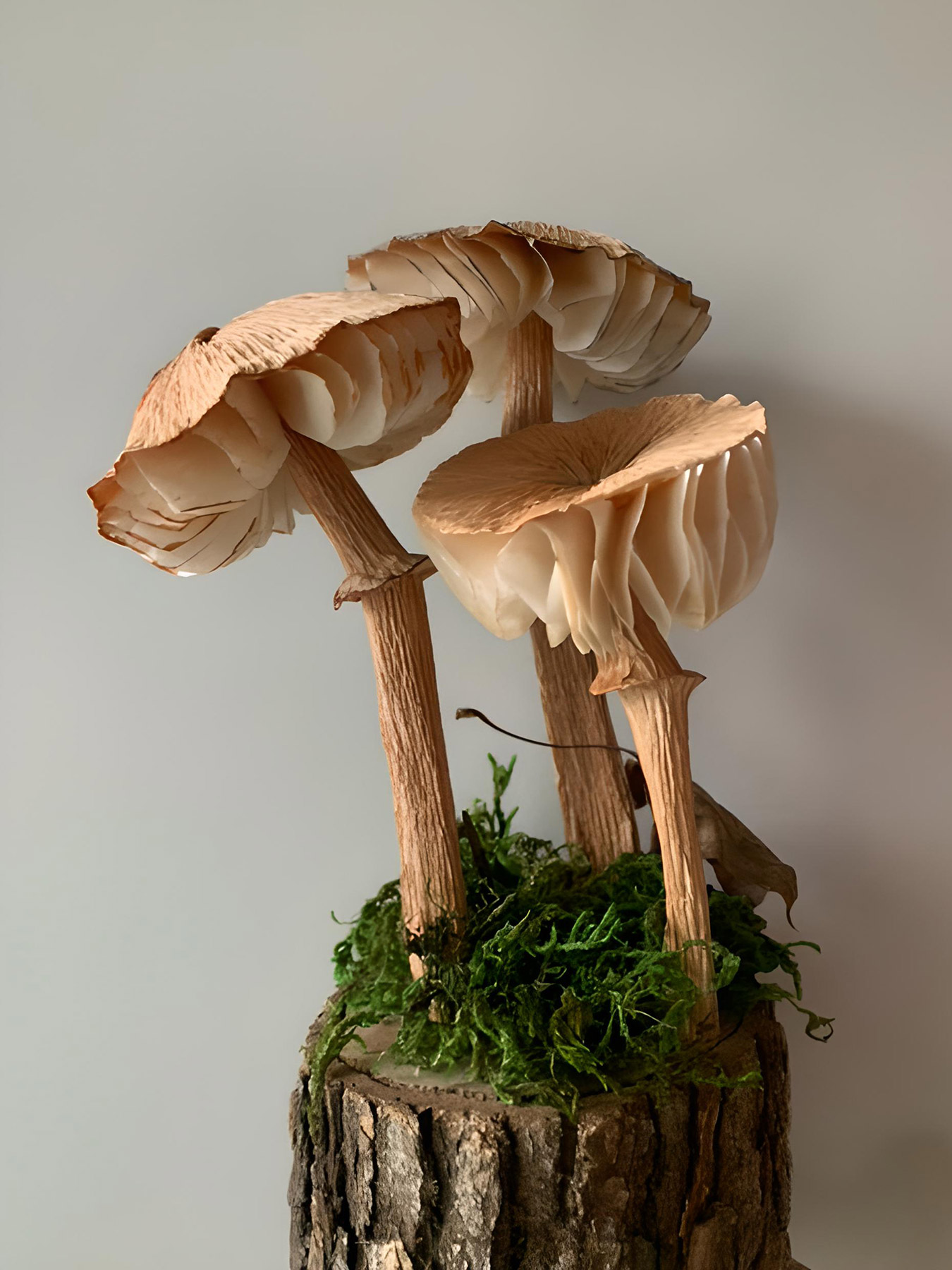 Three tan-colored paper mushrooms arranged together on a log with paper moss against a white background