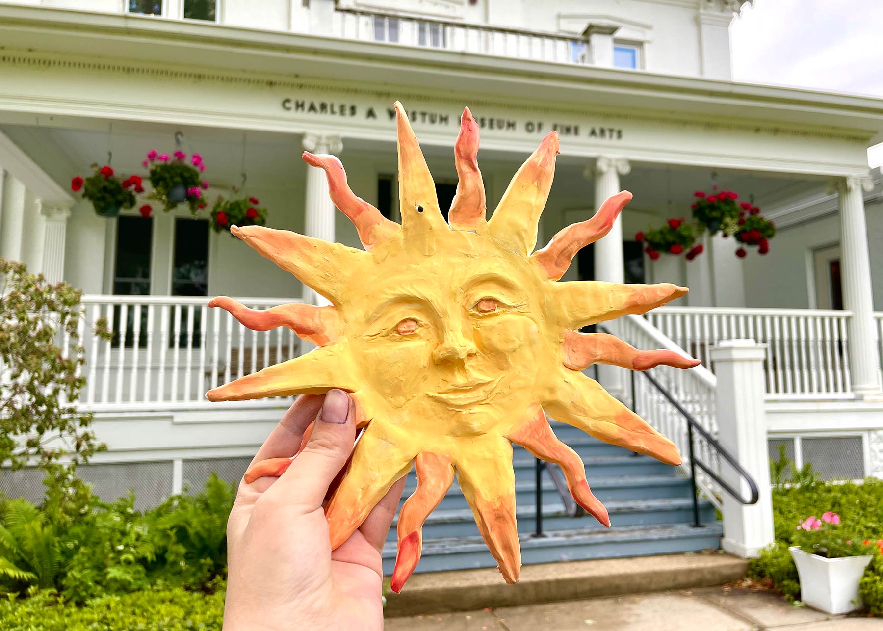 Sun-shaped ceramic object with a face in the center held up in front of RAM's Wustum Museum