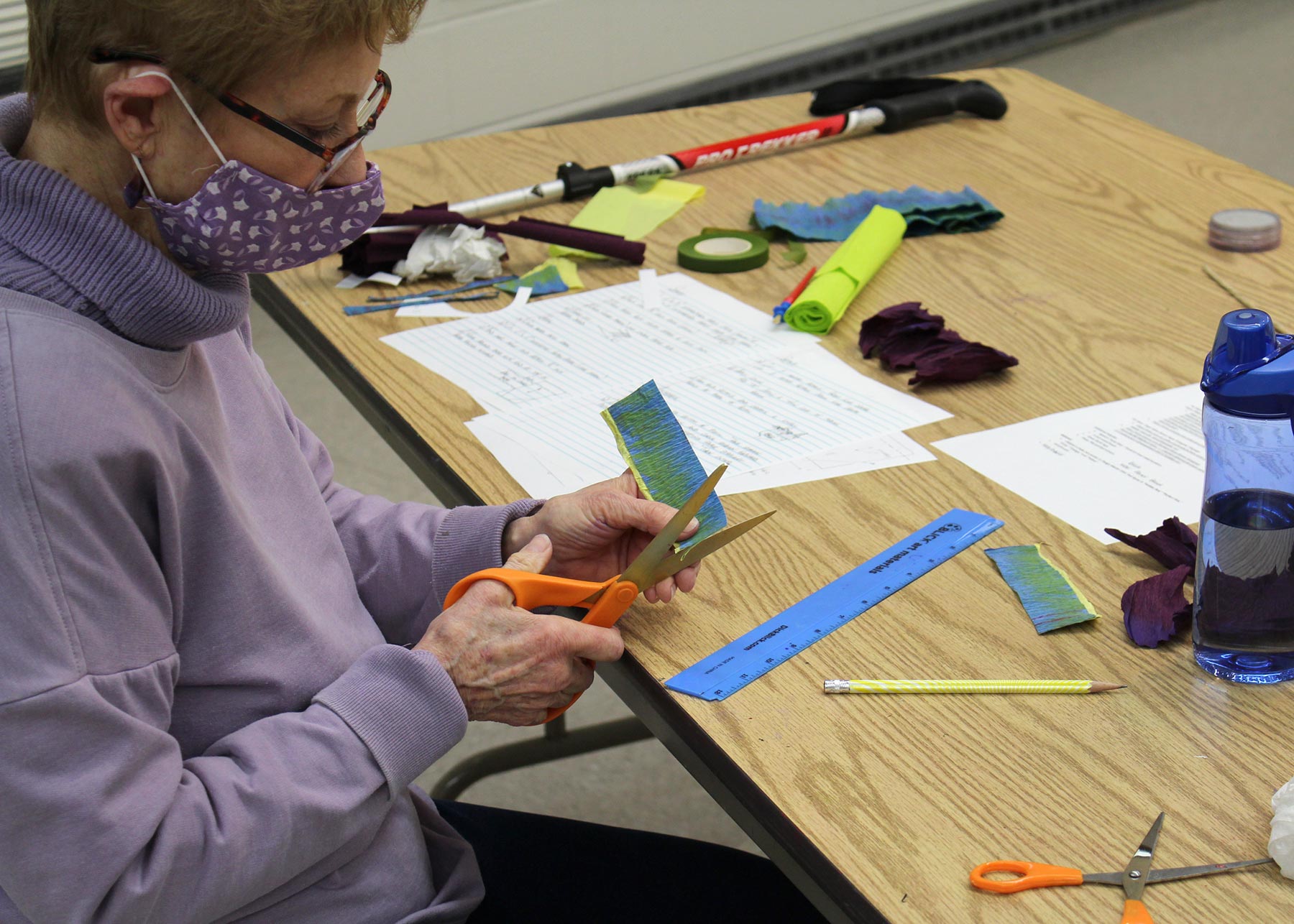 Masked woman sitting at a table covered in art materials, cutting a piece of blue measured paper with orange scissors