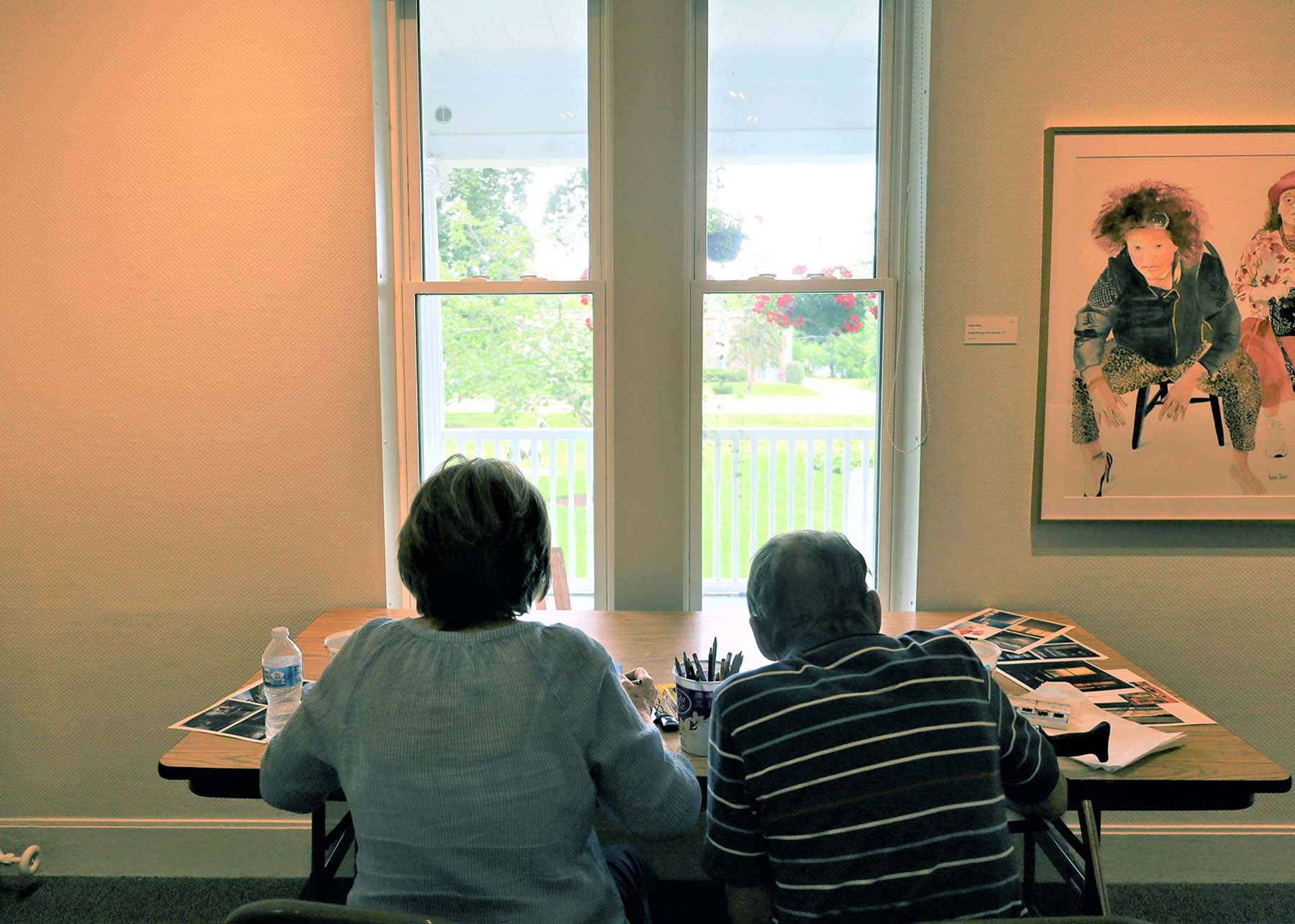 A couple sitting at a table in front of a window inside an art museum gallery, facing away from the viewer. Both people are working on an art project. A garden can be seen from the window.