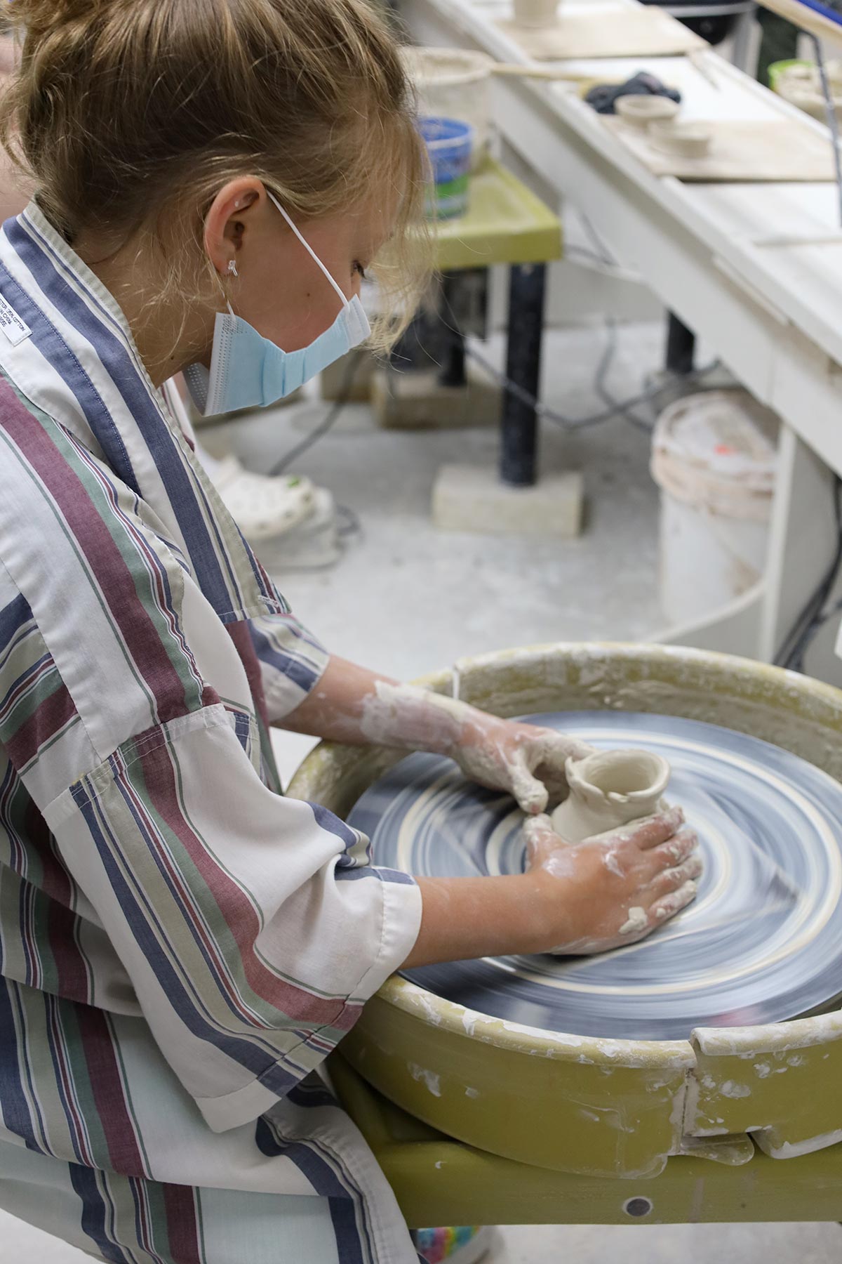 Masked girl wearing a smock while forming a vessel on a potter's wheel