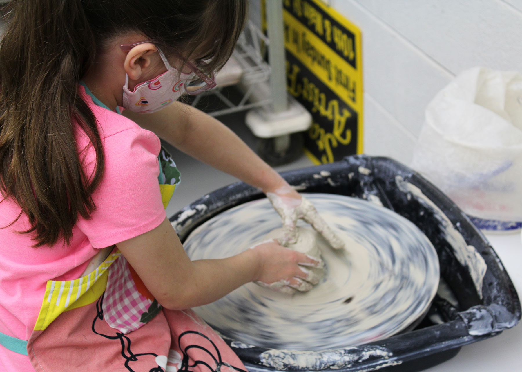 A young child with a pink shirt and face mask is pushing down in a round piece of clay on a potter's wheel.