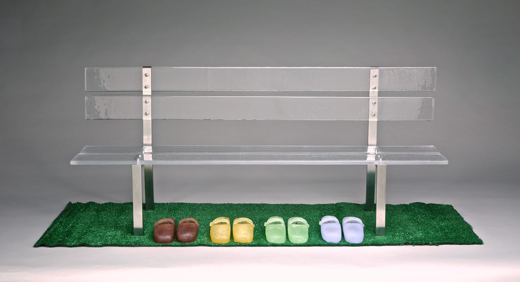 Metal and glass bench and four multicolored pairs of glass shoes atop AstroTurf grass
